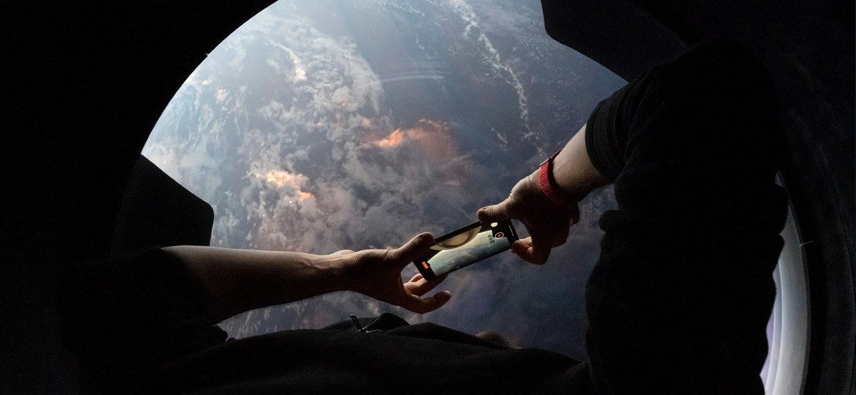 Astronaut's arms holding a device up to a Dragon window with Earth outside