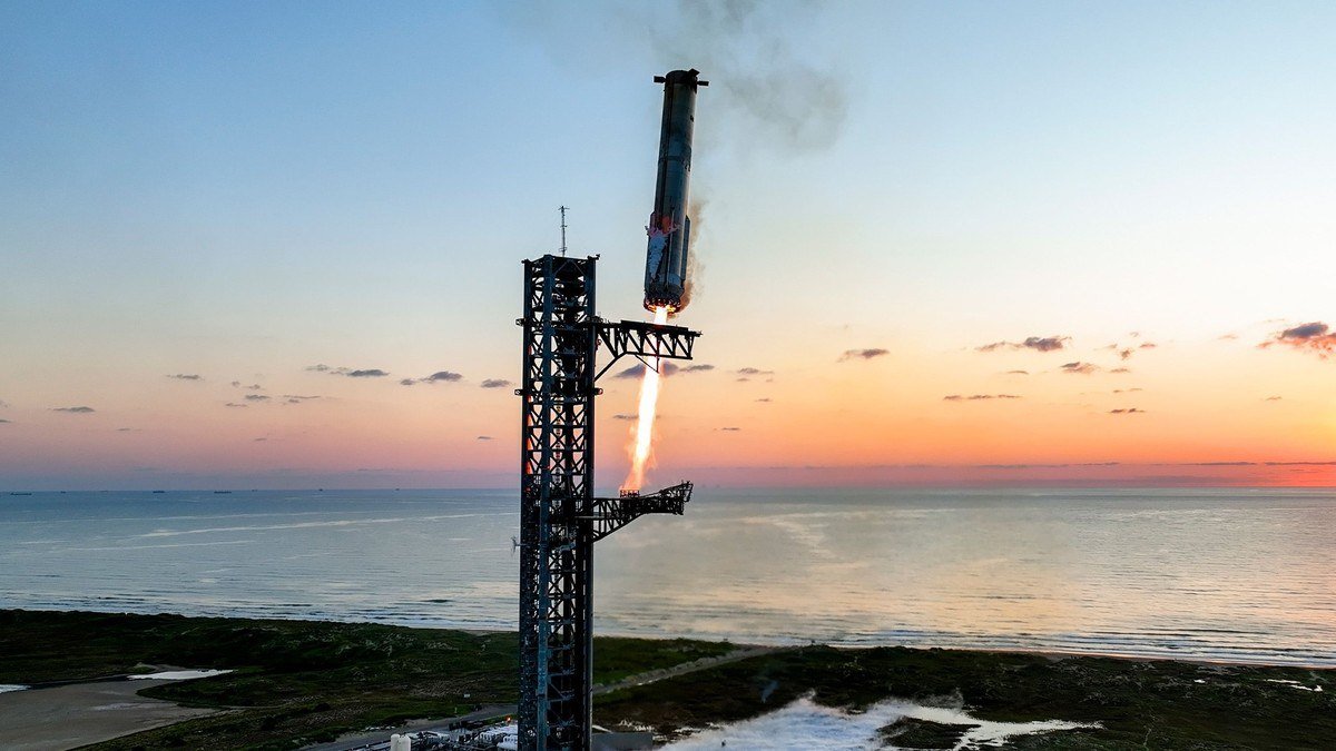 Rocket booster held by a launch tower against a sunset sky over the sea
