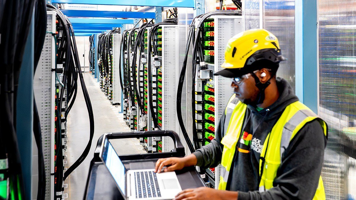 Technician in a yellow hard hat working on a laptop between rows of data center servers