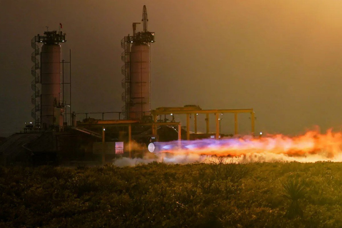 A rocket engine firing a long flame beside two tall test towers in golden evening light