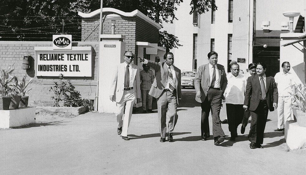 Men in suits walking past the gate of Reliance Textile Industries in a black and white photograph