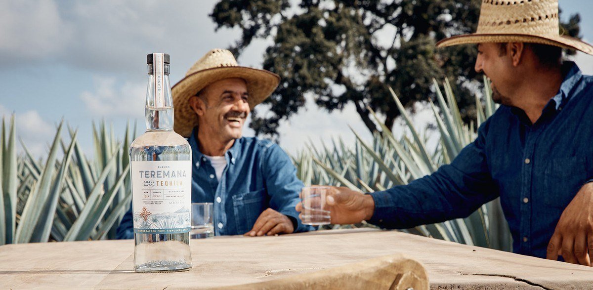 Two men in straw hats at a table among agave plants with a bottle of Teremana tequila