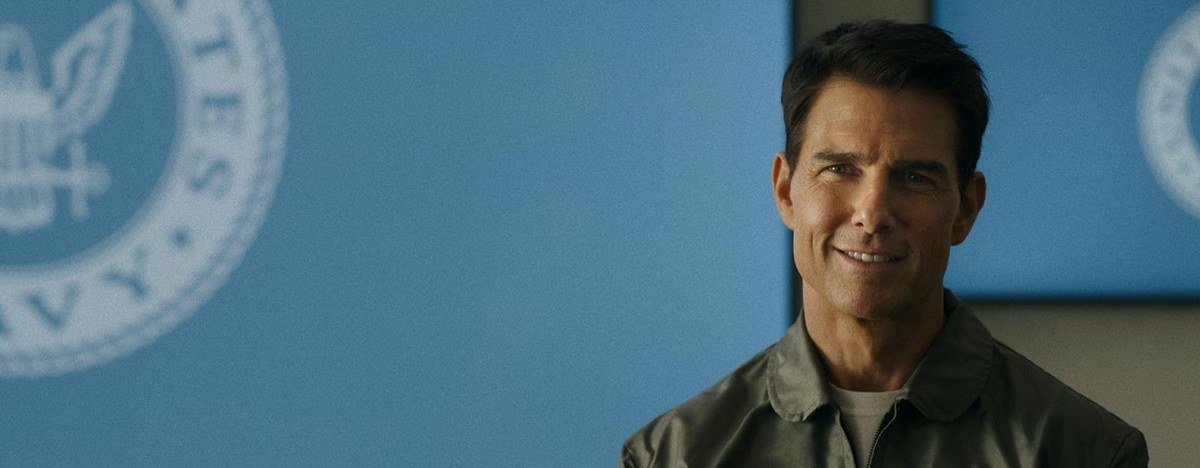 Tom Cruise as Maverick smiling in front of a blue wall carrying a US Navy seal in Top Gun: Maverick