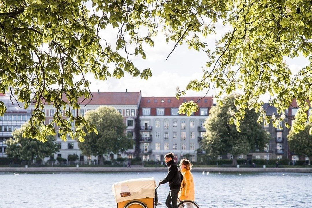 Two people on a cargo bike beside a lake in Copenhagen with apartment buildings behind