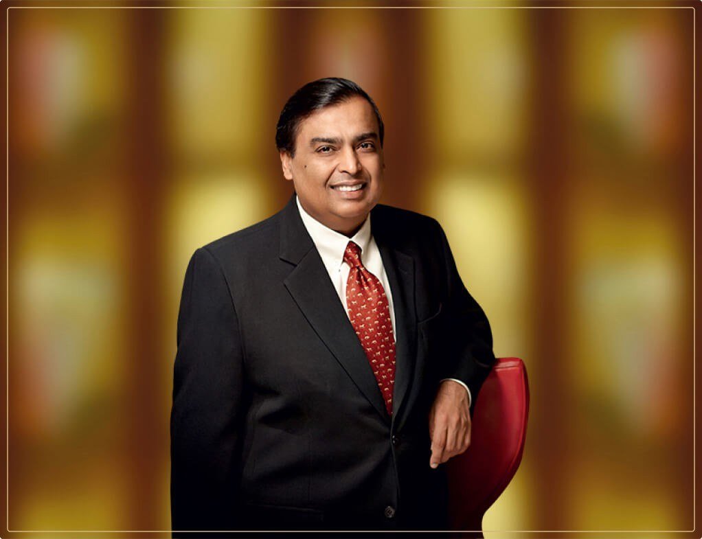 Mukesh Ambani in a dark suit and red patterned tie leaning on a red chair against a blurred gold background