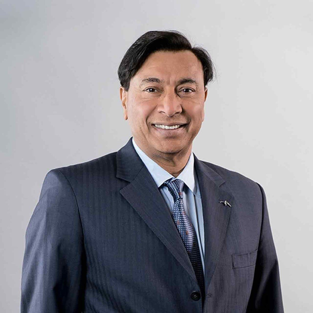 Studio portrait of Lakshmi Mittal, smiling in a navy suit and blue tie against a light grey background