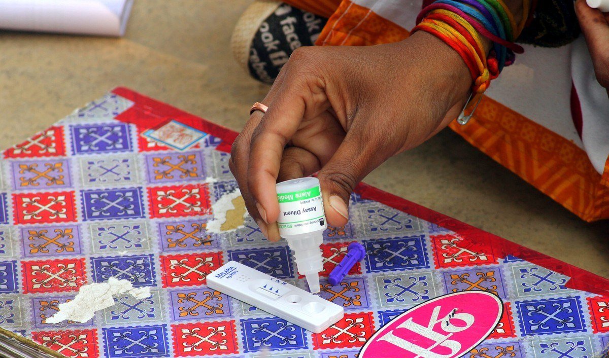 A hand pouring buffer solution from a dropper bottle onto a rapid diagnostic test kit in a Sun Pharma health programme